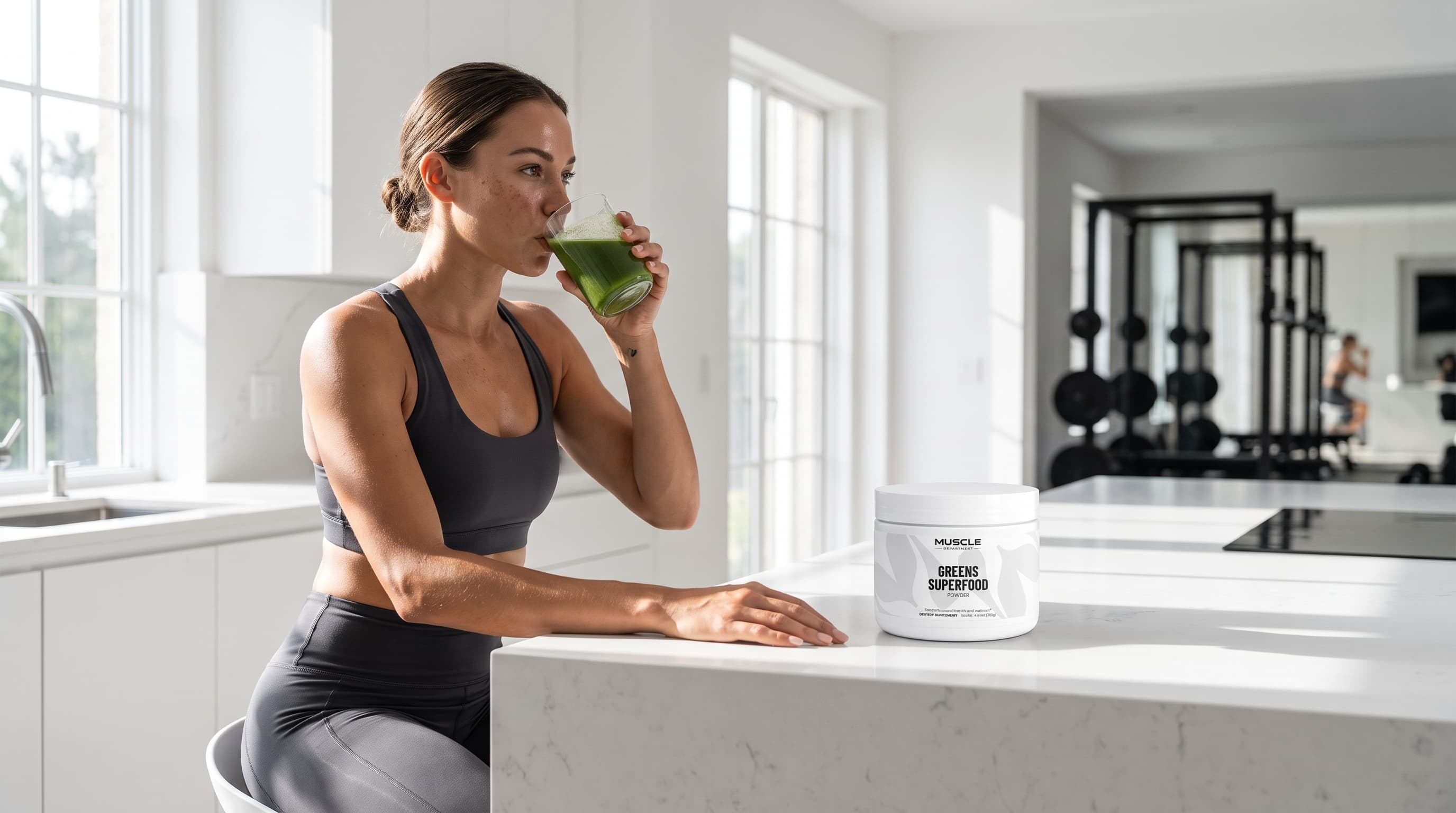 A woman in a black sports bra and leggings sits at a white kitchen island, drinking from a green cup while a white tub of Muscle Department Greens Superfood Powder rests on the counter in a bright, modern kitchen.