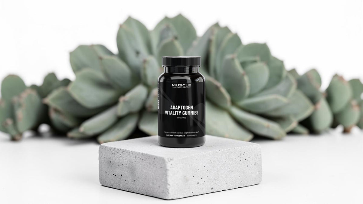 A black bottle of Muscle Department Adaptogen Vitality Gummies sits on a light gray concrete block, with blurred green succulent plants in the background against a white studio backdrop.